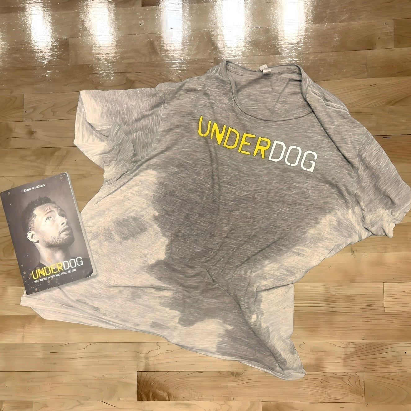Gray t-shirt with 'UNDERDOG' text and a book titled 'UNDERDOG' on a wooden floor.